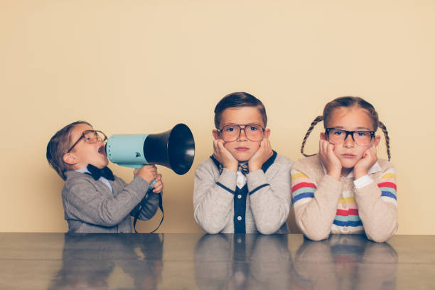 A young boy in glasses and a nerd outfit is shouting at his siblings through a megaphone. The big brother and big sister are looking at the camera with an annoyed look on their faces while plugging their ears. The little brother is bossing his older siblings around.